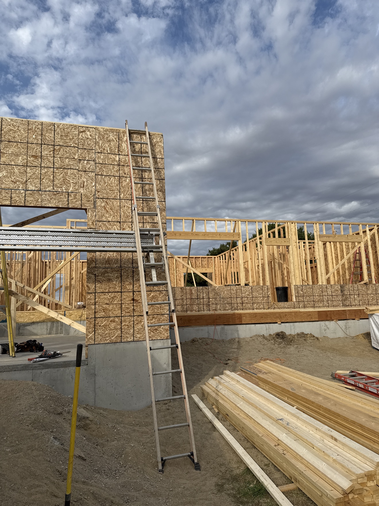 Current house framing project with sheathed exterior wall, wood framing, ladders, and stacked lumber